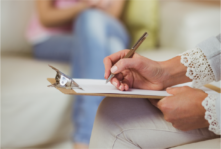 Profissional da saúde mental fazendo anotações durante sessão com paciente ao fundo.
